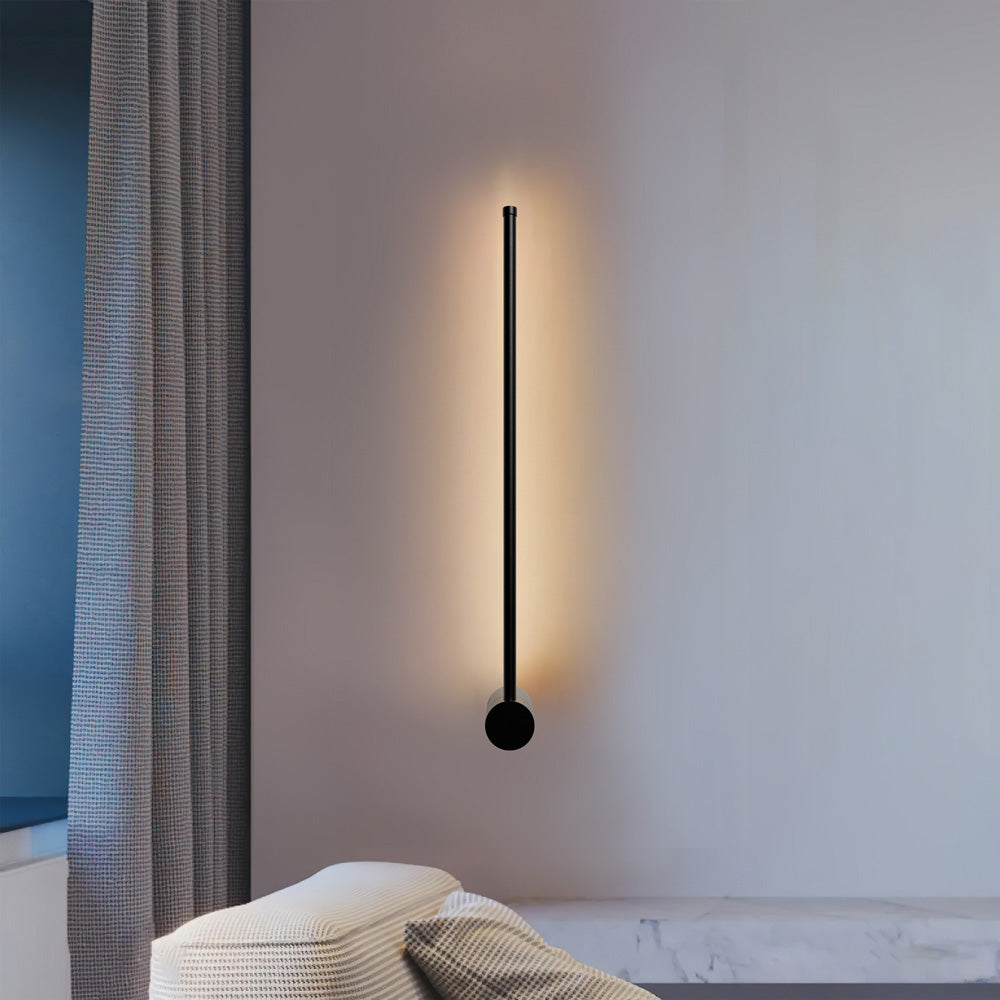 Modern black pila rotatable linear LED wall sconce mounted vertically, casting a warm glow in a minimalist living space