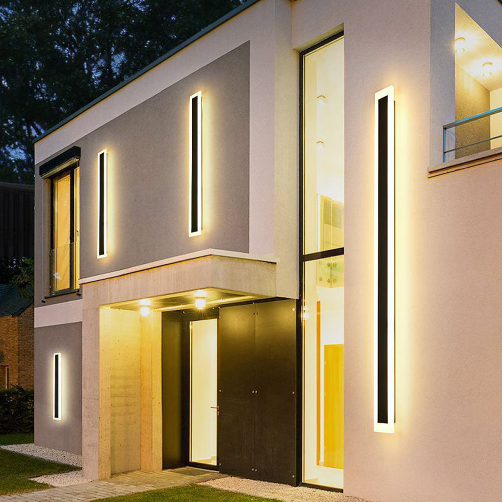 Three long black exterior xura wall lamps installed vertically on the pale facade of a contemporary house